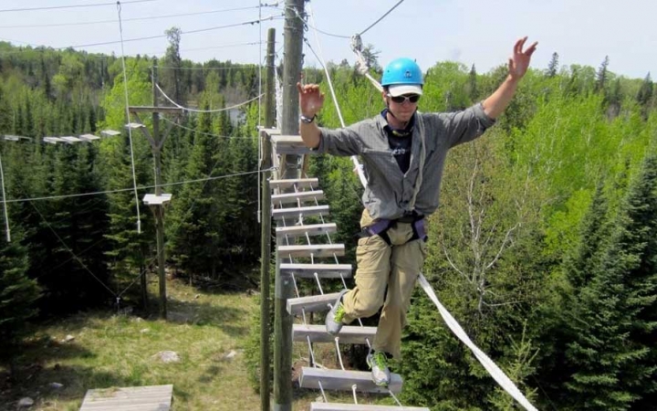 A person wearing safety gear and secured by ropes balances on an obstacle on a high ropes course. 